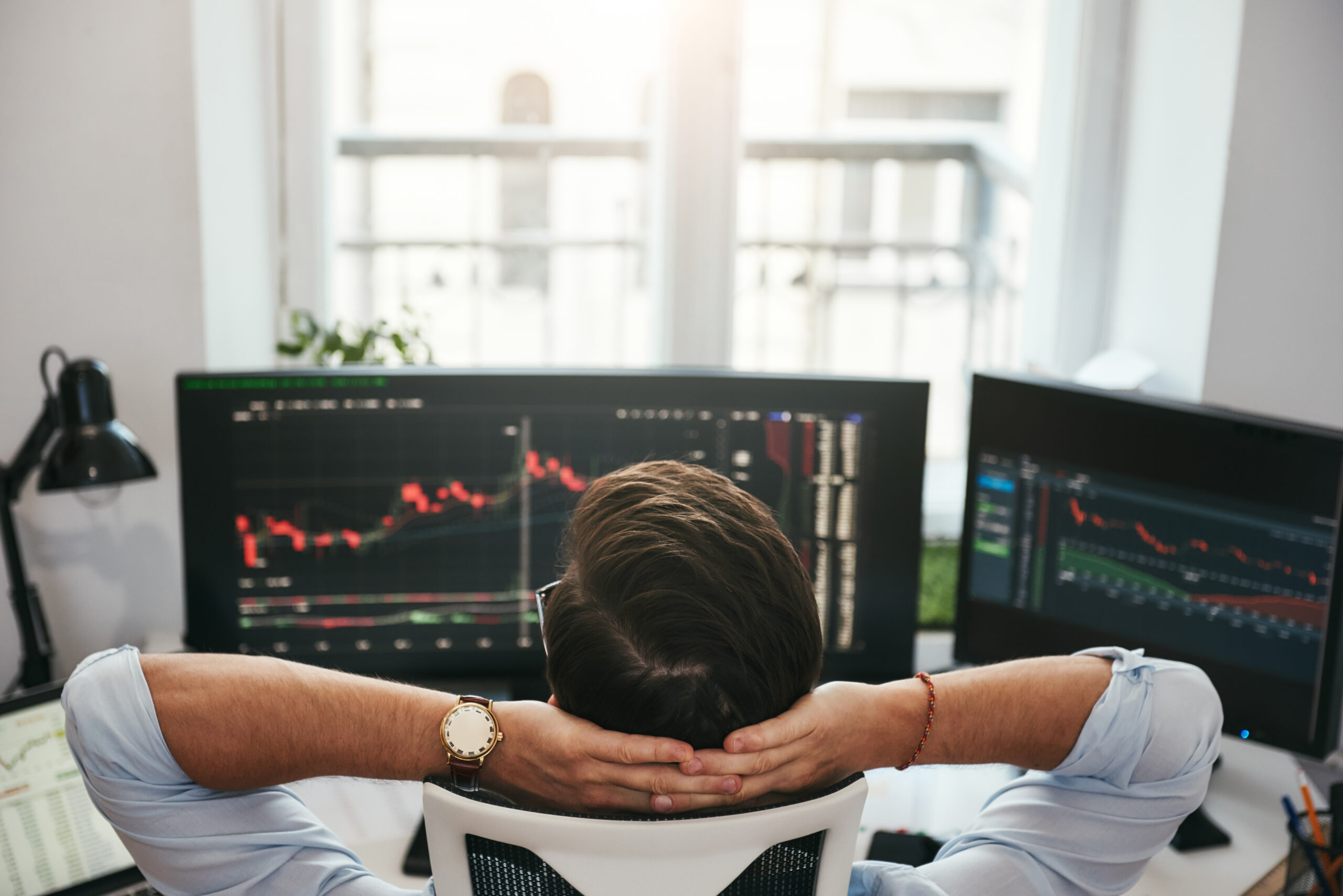 Time to relax. Back view of young businessman or trader holding hands behind head while looking at charts and financial data on computers in his modern office. Forex market. Trade concept. Investment concept